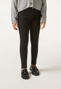 Jeans skinny negros con una textura suave, cintura alta y costuras laterales, combinados con zapatos negros de slip-on con un diseño texturizado.