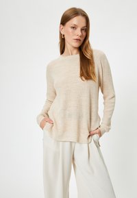 Ljust beige stickad tröja med avslappnad passform, långa ärmar och fickor. Tyget har en subtil ribbad struktur och en mjuk yta.