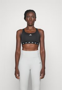 Sostén deportivo negro con logotipo blanco en el centro y patrones triangulares en el borde inferior. Combinado con mallas de cintura alta en gris claro.