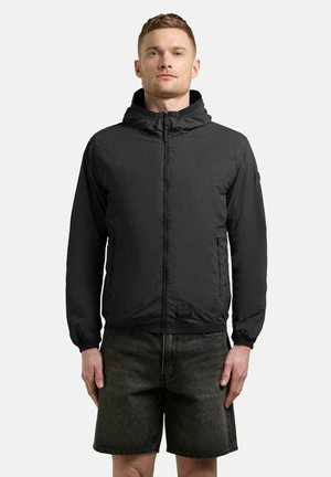 Mand iført en sort hættetrøje med lynlås og mørke denimshorts, stående med armene afslappet ned langs siden mod en ensartet lys baggrund.