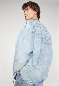 Kurtka denimowa w kolorze jasnoniebieskim z efektem przetarcia, w oversize'owym kroju, z kontrastowym stębnowaniem i charakterystycznym fasonem pleców z zakrzywionymi liniami.