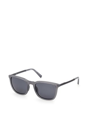 Graue rechteckige Sonnenbrille mit dunkel getönten Gläsern und dünnen schwarzen Bügeln, leicht schräg auf weißem Hintergrund gezeigt.