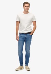 Camiseta blanca de cuello redondo, vaqueros ajustados de color azul claro, zapatillas beige, cinturón negro y reloj plateado. Textura suave en todo.
