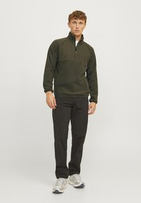 Dark green fleece pullover with a half-zip collar, featuring a textured upper panel. Paired with black pants and light-colored sneakers.