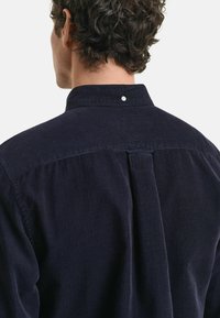 Mörkblå corduroyskjorta med högt krage, vertikal ribbad textur, och en enda knappdetalj på baksidan.