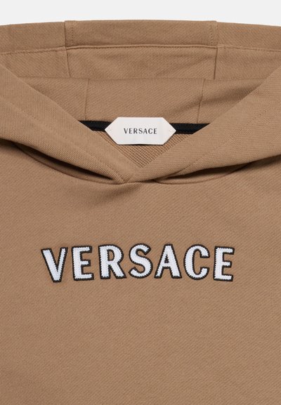 Μπεζ φούτερ με κουκούλα, με τη λέξη «VERSACE» κεντημένη με μεγάλα λευκά γράμματα περιγραμμένα με μαύρο στο στήθος και μια λευκή ετικέτα Versace στο εσωτερικό του γιακά.