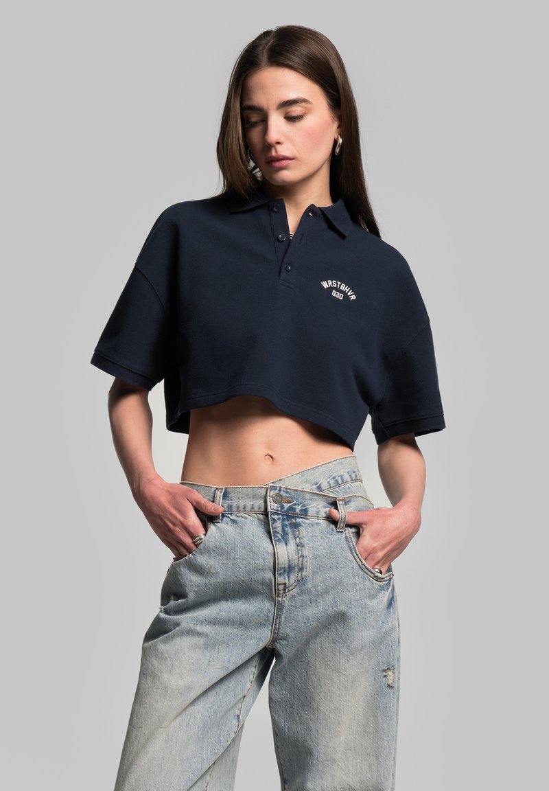 Granatowa, skrócona koszulka polo z krótkim rękawem i zapięciem na guziki; posiada białe logo tekstowe na piersi. Uzupełniona o wyblakłe jasnoniebieskie dżinsy.