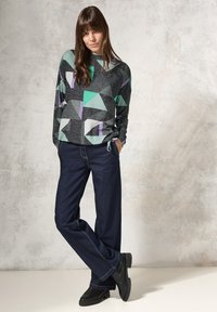 De trui heeft een geometrisch patroon in zwart, groen en paars, met lange mouwen. Gecombineerd met donkerblauwe wijde jeans en zwarte schoenen.