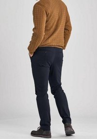 Man i brun kabelstickad tröja, mörkblå byxor och mörkbruna skor står med händerna i fickorna, vänd bort.