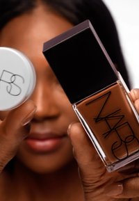Flacon de fond de teint NARS avec un bouchon noir carré, une base en verre transparent et un liquide brun. Accompagné d'un récipient rond blanc avec un logo noir.