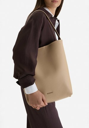 Mujer con ropa oscura sosteniendo un bolso beige con textura, asa corta y pequeño logo dorado de la marca cerca de la parte inferior.