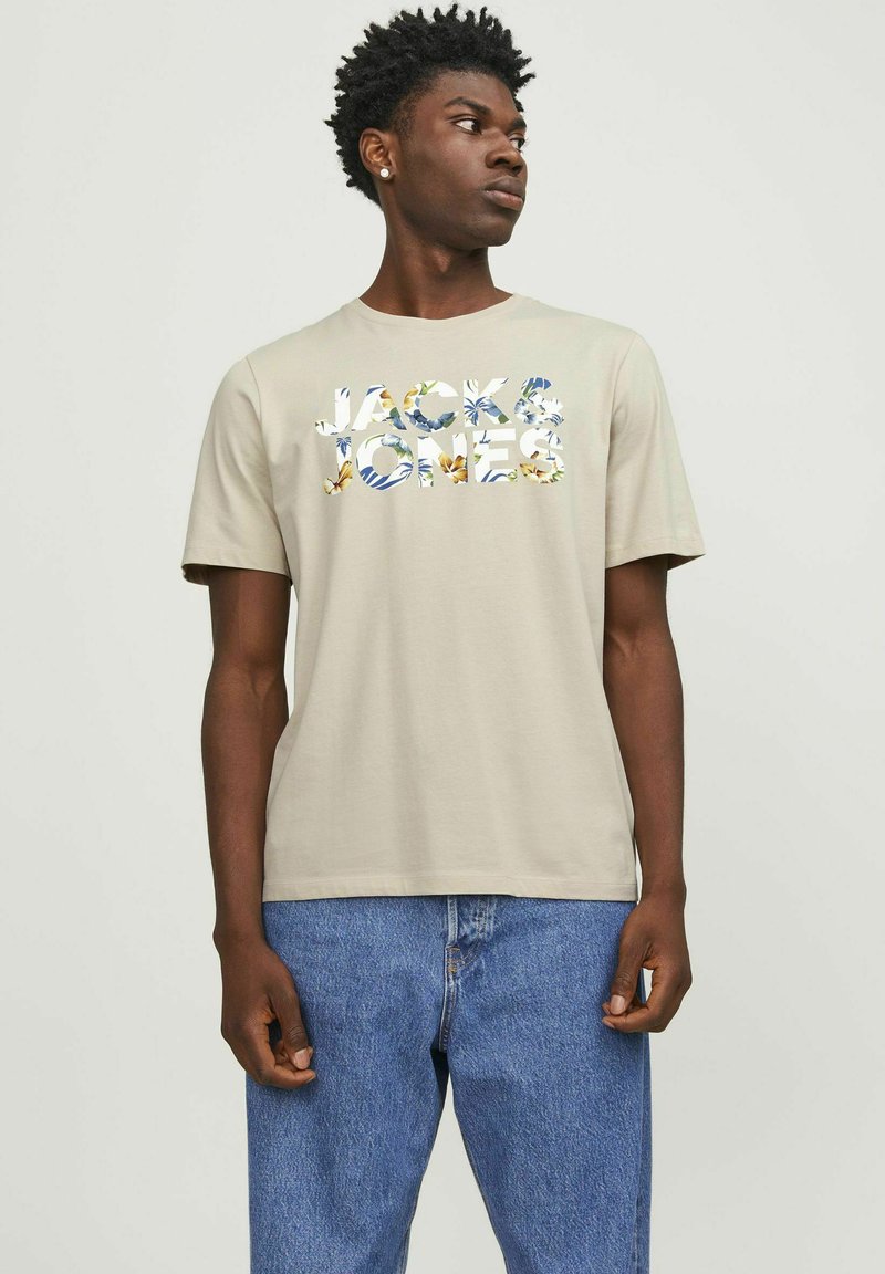 Beige bomull t-skjorte med blomsterdesign og teksten "JACK & JONES." Korte ermer, rund hals, sammen med blå jeans.