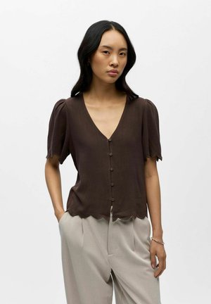 Žena s dlhými čiernymi vlasmi, oblečená v hnedej blúzke s gombíkmi, výstrihom do V a vlnkovanými okrajmi, a svetlosivých širokých nohaviciach.
