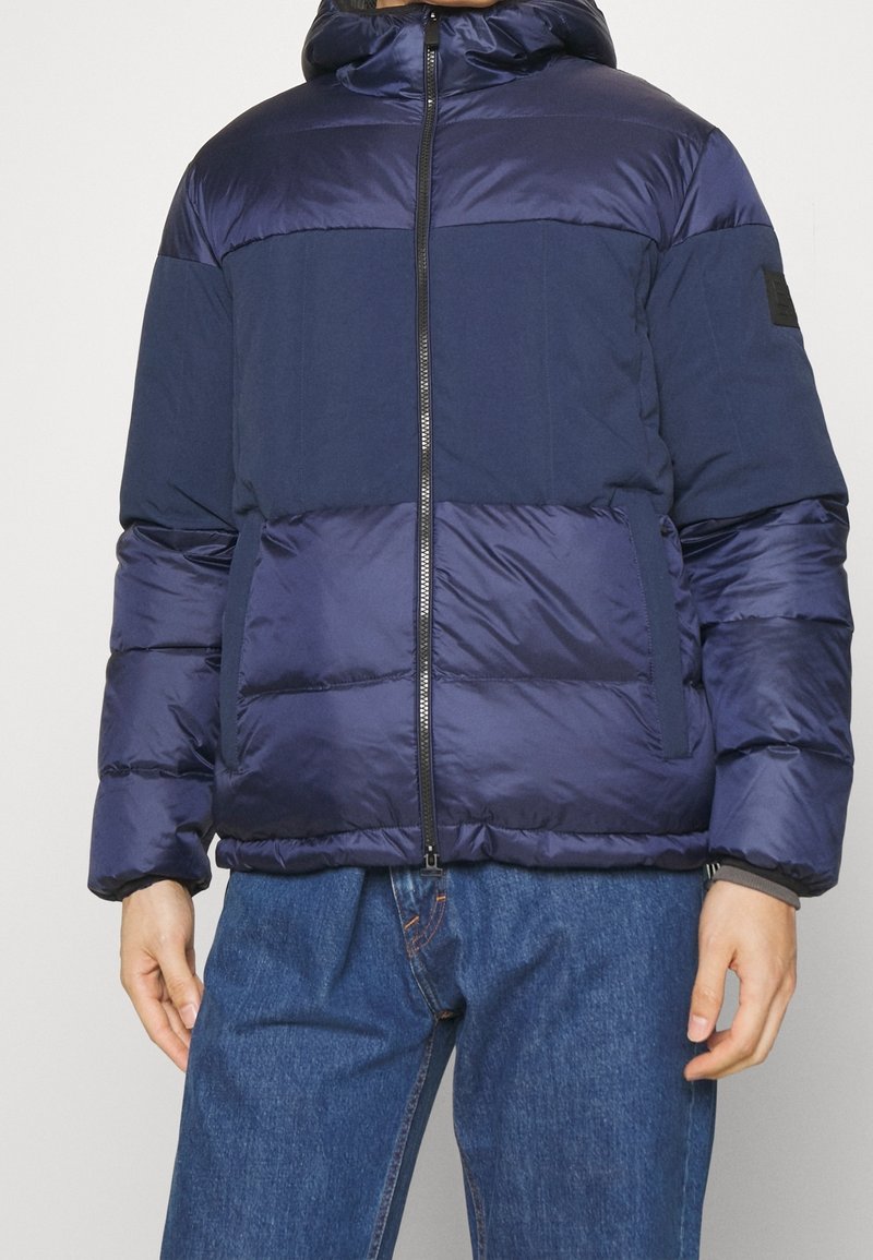Granatowa puchowa kurtka zapinana na zamek, z kapturem i kontrastową fakturą; górna część gładka, dolna pikowana, z kieszeniami po bokach.