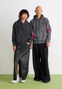 Zwei Personen tragen dunkelgraue Kapuzenpullis mit Fronttaschen und Reißverschlüssen. Das Outfit wird mit schwarzen Hosen und einem Ledrock kombiniert.