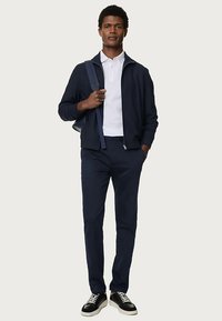 Marineblauwe jas met rits over een wit poloshirt, gecombineerd met marineblauwe broek en zwarte sneakers. Glad materiaal, ontspannen pasvorm en subtiele designdetails.