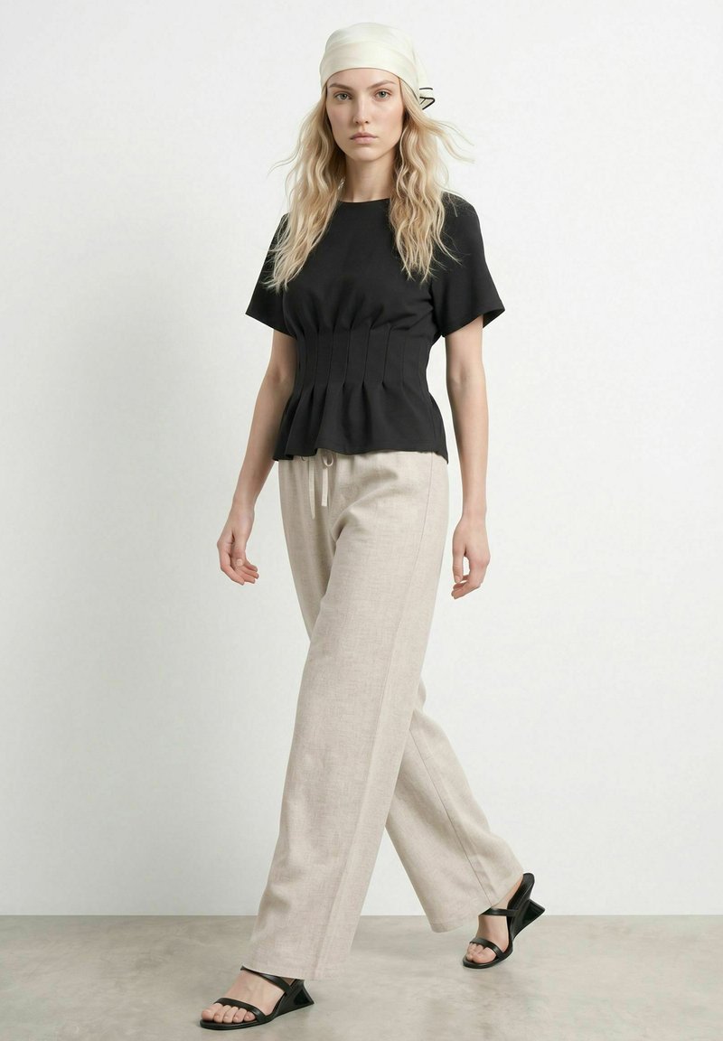 Femme marchant portant un haut noir à manches courtes, un pantalon beige à jambes larges, des sandales noires et un foulard clair sur un fond uni.
