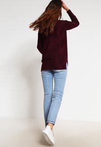 Burgundy stickad tröja med långa ärmar, sidofickor, i kombination med ljusblå skinny jeans och vita sneakers. Bakifrån, vågigt hår synligt.