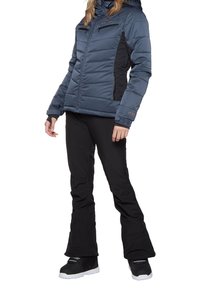 Eine Frau trägt eine blaue gepolsterte Jacke, schwarze Schneehosen und schwarze Schneestiefel und steht vor einem weißen Hintergrund.