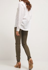 Camisa blanca de manga larga combinada con pantalones ajustados verdes oliva y sandalias de tacón beige. La camisa presenta un diseño informal y costuras visibles.
