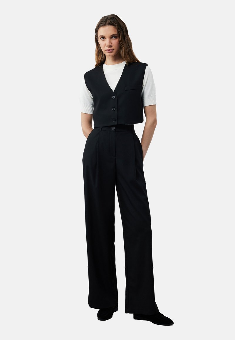 Zwarte cropped top met een V-hals en knopen, gecombineerd met hoge zwarte wijde broek. Witte korte mouw shirt eronder.