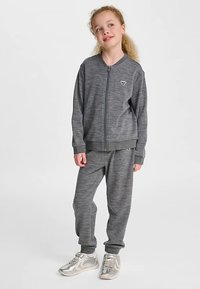Grå zip-up jacka och matchande mjukisbyxor i mjuk textur, med en liten logotyp. Matchas med silverfärgade träningsskor.
