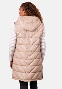 Woman with curly hair wearing a beige quilted sleeveless hooded vest over a white and beige striped long-sleeve shirt, facing away.