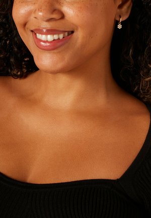 Boucles d'oreilles créoles en or avec un motif floral ornées de petites pierres claires. Le modèle a les cheveux bouclés et porte un haut noir.