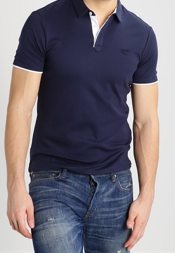 Marinblå polo med vit krage och manschetter, slim fit-design, med en diskret logotyp på vänster bröst. Kombinerad med slitna jeans.