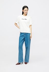 Μπεζ βαμβακερό t-shirt με κεντητό λογότυπο "Tommy Hilfiger", κοντά μανίκια και κουμπιά στον ώμο, συνδυασμένο με φαρδιά μπλε τζιν παντελόνια.