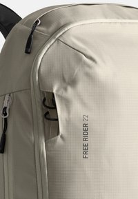 Beige Stoff-Rucksack mit glatter Textur, Reißverschlussfächern und einem seitlichen elastischen Kordelzug für externe Aufbewahrung. Das Etikett trägt die Aufschrift "FREE RIDER 22."