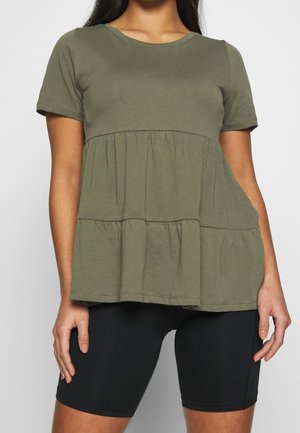 Kobieta ubrana w oliwkowo-zieloną, krótką, warstwową bluzkę i czarne dopasowane szorty do kolan, stojąca na tle jasnoszarego tła.