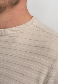 Beige gebreide trui met textuur en horizontale strepen, met een geribde ronde halslijn en zachte stof. Close-up van het schoudergebied.