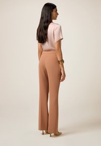 Haut rose clair à manches courtes associé à un pantalon large beige taille haute ; tissu doux avec un léger éclat ; le modèle porte un bracelet décoratif.