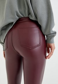 Person som bär högmidjade, burgundyröda imitationläderbyxor och en lös, långärmad grå topp, stående mot en enkel bakgrund.