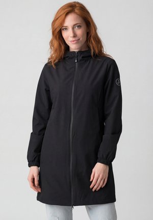 Frau mit roten Haaren, die eine lange schwarze Jacke mit Reißverschluss und elastischen Bündchen sowie hellblaue Jeans trägt, steht vor einem schlichten grauen Hintergrund.