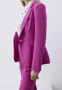 Blazer fuchsia sur mesure en tissu lisse, avec un col à revers cranté, une fermeture à un bouton et des poches latérales, porté avec un chemisier blanc.