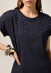 Camiseta de color azul marino con gemas decorativas negras y azules dispersas en la parte delantera, mangas cortas y cuello redondo. Incluye dos pulseras.