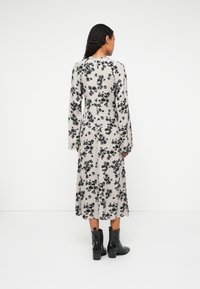 Robe longue à motifs floraux en beige et noir, avec des manches longues et une taille cintrée. Portée avec des bottines noires à la cheville. Vue de dos.