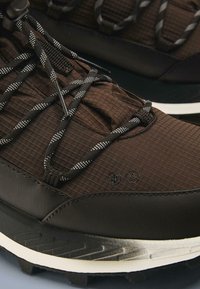 Pantofi de drumeție maro cu partea superioară texturată, șireturi alb-negru, talpă de cauciuc și accente duble de branding.