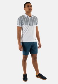 Polo bianco con accenti a motivo blu nella parte superiore. Indossato con pantaloni corti blu e scarpe da ginnastica nere con strisce arancioni.