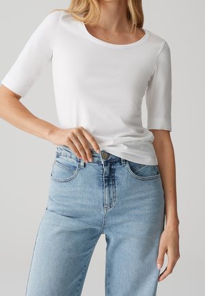 Femme portant un t-shirt blanc uni à manches courtes et un jean bleu clair taille haute, main reposant près de la ceinture.