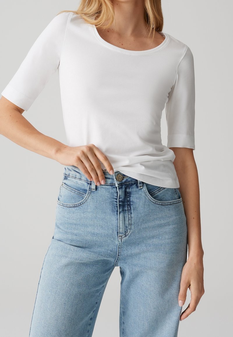 Femme portant un t-shirt blanc uni à manches courtes et un jean bleu clair taille haute, main reposant près de la ceinture.