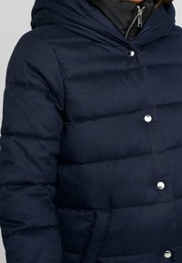 Marinblå pufferjacka med huva, vadderad design och metallknappar. Har en slät yta och quiltat mönster.