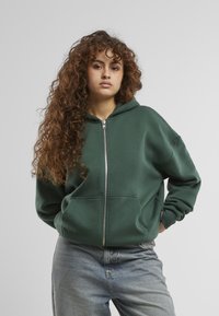 Grøn zip-up hoodie i blødt materiale med hætte, frontlommer og ribkant. Bæres med lyseblå jeans.