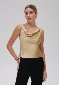 Top de satén con cuello de cowl en dorado claro, que presenta una textura suave y un diseño sin mangas, combinado con pantalones negros.