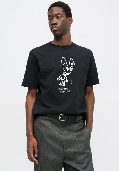 Muž v černém tričku Maison Kitsuné s bílým abstraktním designem lišky a postavy, v kombinaci se šedými kalhotami a černým páskem, ruka v kapse.