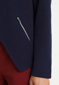 Chaqueta azul marino con diseño elegante, tejido texturizado y un bolsillo con cremallera plateada en el lado izquierdo, combinada con pantalones rojos.