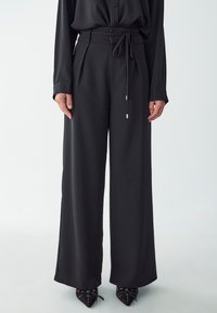 Black wide-leg trousers with a tied waist, pleated front, and smooth texture. Worn with pointed black shoes featuring gold accents.