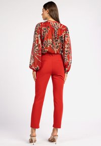 Pantalons rouges avec une taille ajustée et des jambes droites, associés à un blouse transparent à motif floral avec des manches bouffantes.